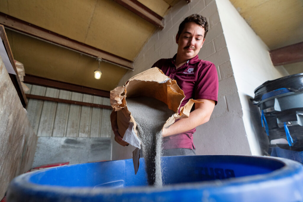 Guy pouring in concrete into a blue barrel wearing a Texas A&M shirt. 