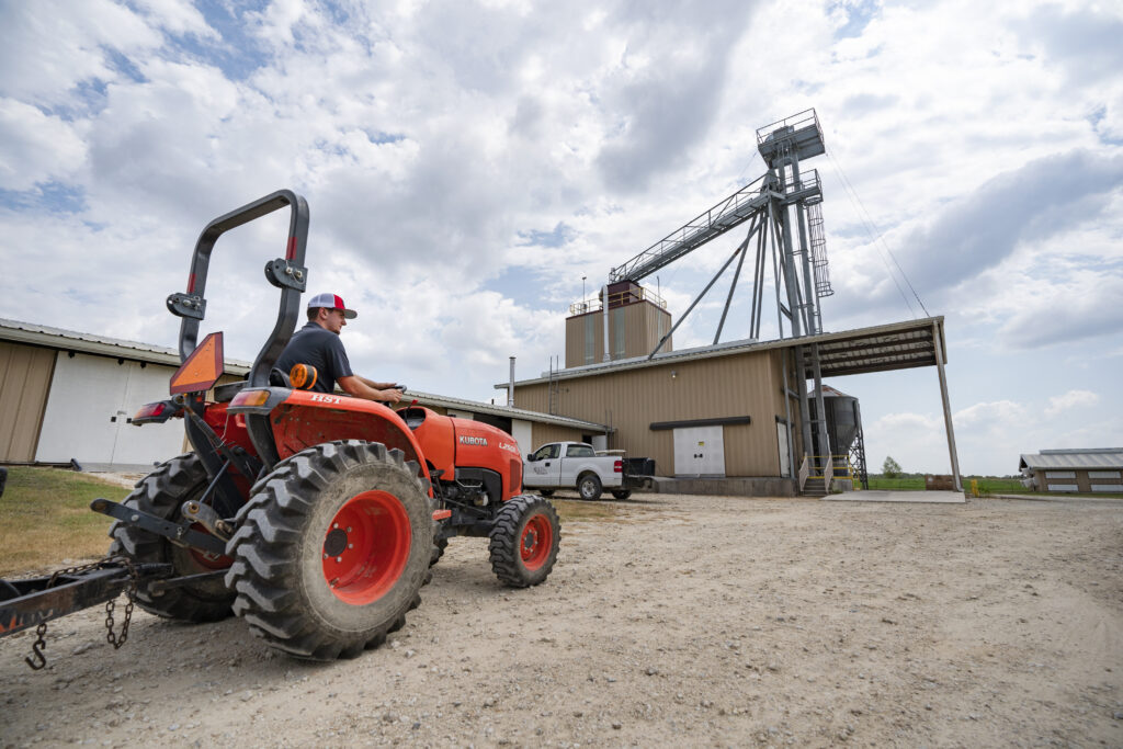 A man operates a tractor on a dirt road, surrounded by greenery and open fields, showcasing rural life and agriculture.
