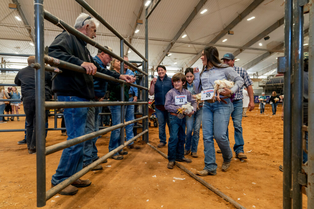 A group of people, including children holding chickens and wearing numbered tags, walk toward a livestock pen inside a large indoor arena with dirt flooring.